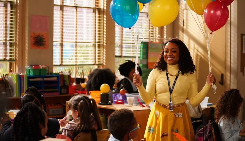 Quinta Brunson como Janine em uma sala de aula segurando balões coloridos, sorri e veste roupas amarelas