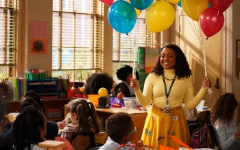 Quinta Brunson como Janine em uma sala de aula segurando balões coloridos, sorri e veste roupas amarelas