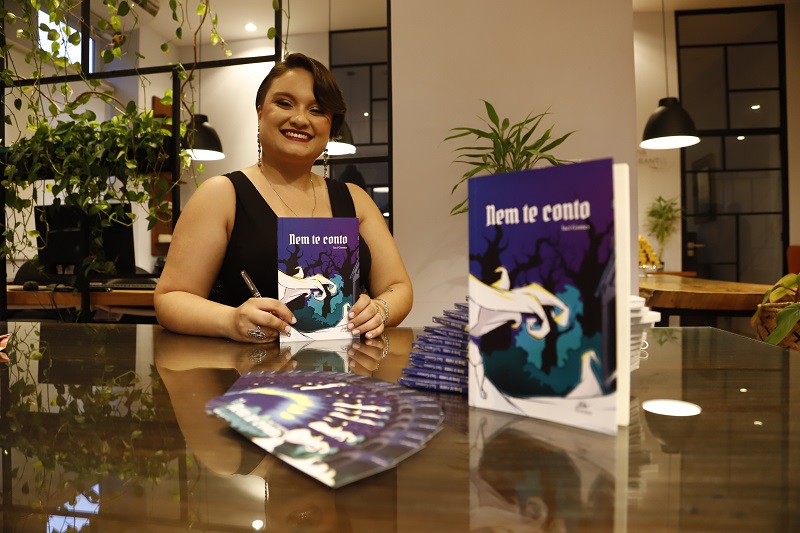 A autora Iaci Gomes está sentada e segurando um exemplar da edição do livro "Nem te Conto". Ao redor dela há o reflexo de luzes e plantas decorando o ambiente. 
