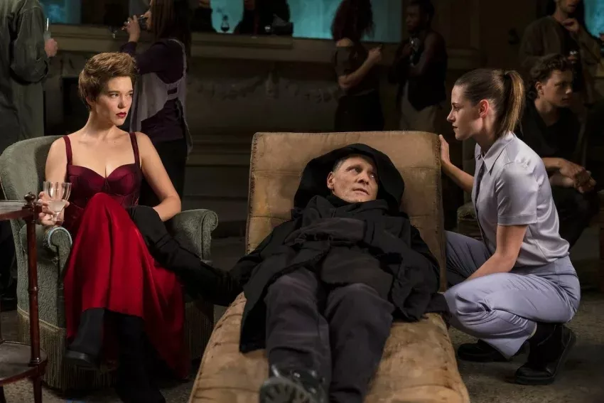 Viggo Mortensen deitado no sofá com uma roupa preta, Léa Seydoux, na lateral esquerda, sentada, com um vestido vermelho e segurando uma taça, e Kristen Stewart , na lateral direita, agachada e com uma roupa azul clara. Cena do filme "Crimes do Futuro" (2022), de Cronenberg.