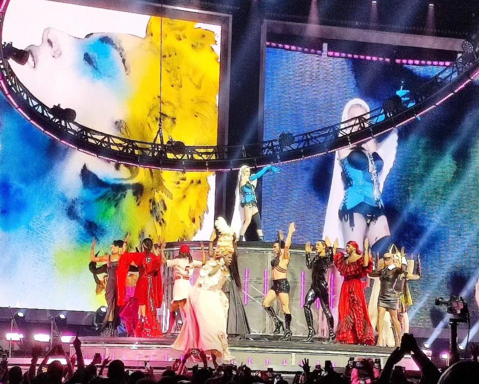 Madonna canta sobre uma estrutura elevada enquanto os dançarinos giram en torno, vestidos com figurinos das várias fases da cantora.