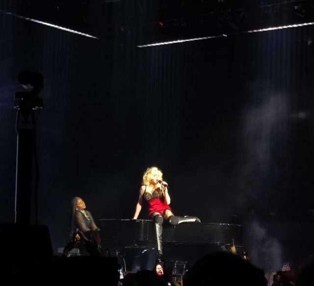 Madonna vestida com um baby doll vermelho canta sentada sobre um piano, com a filha Mercy James vestida com um smoking preto tocando o instrumento. 