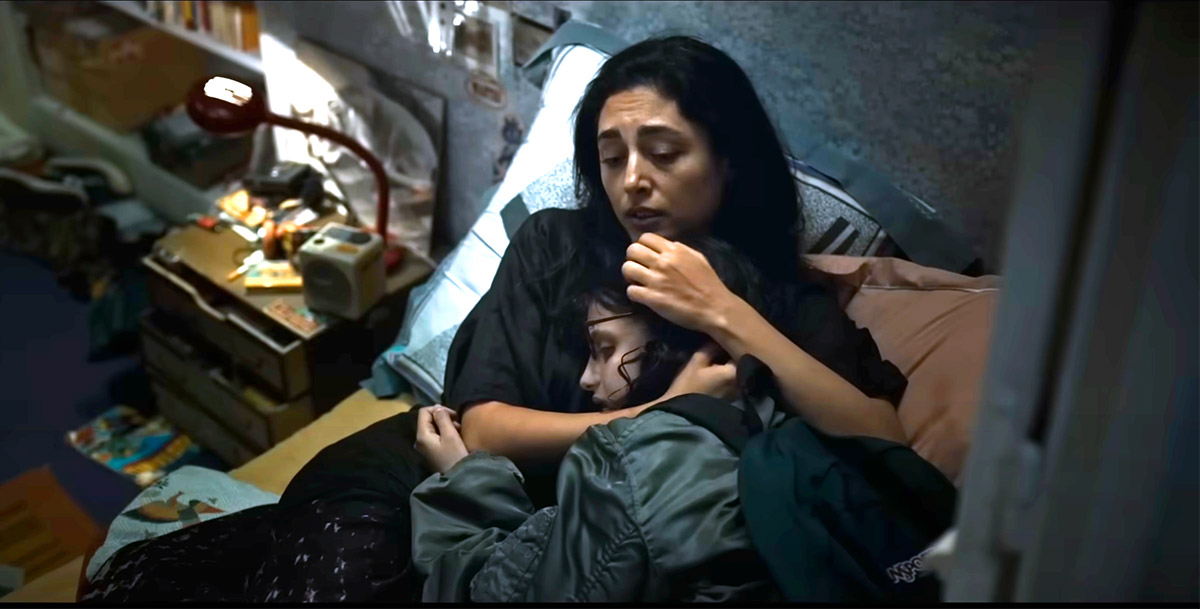 Cena com duas pessoas em uma cama, uma mulher e uma criança abraçada a ela, em ambiente de quarto.
