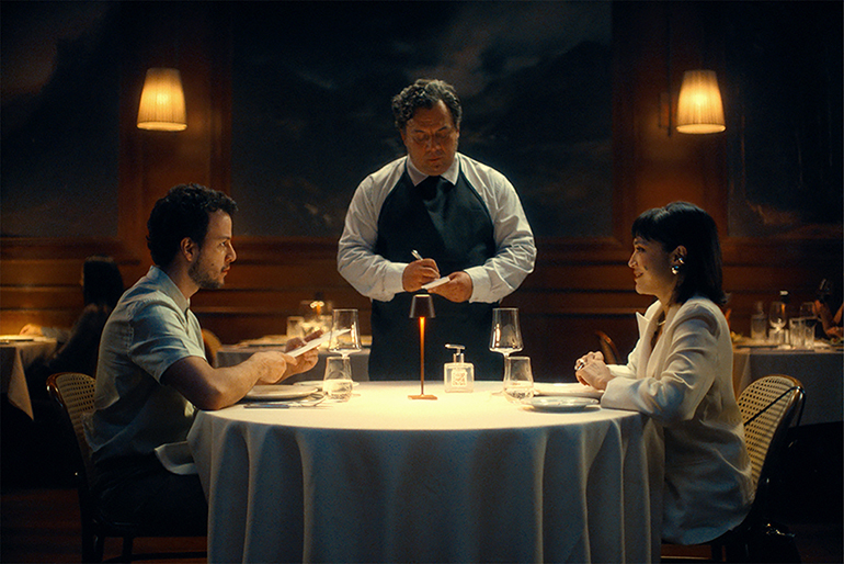Cena de um filme de suspense com um garçom entregando o cartão na mesa de um casal em um restaurante sofisticado, com iluminação suave e ambiente elegante.