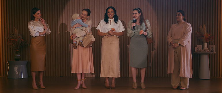 Cena de uma apresentação ou evento com várias mulheres segurando microfone e um bebê, todas vestidas com roupas em tons pastéis em um cenário com iluminação suave.