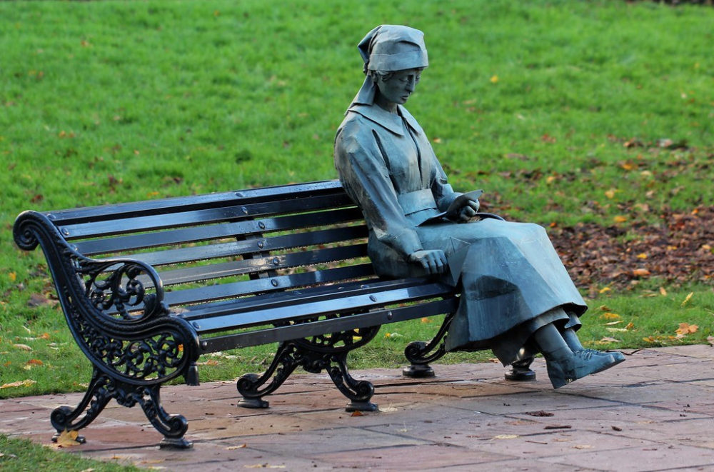 Estátua de uma mulher sentada em um banco de parque, usando chapéu e lendo um livro, em um ambiente com gramado verde ao fundo.