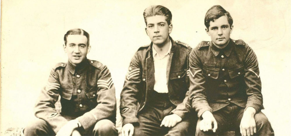 Foto antiga de três jovens soldados uniformizados, sentados em uma pose casual, com expressão séria.