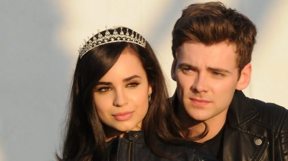 Cena de um casal jovem, uma mulher com tiara e cabelo escuro, e um homem com cabelo castanho, ambos vestindo jaquetas de couro, em uma expressão pensativa ao ar livre.