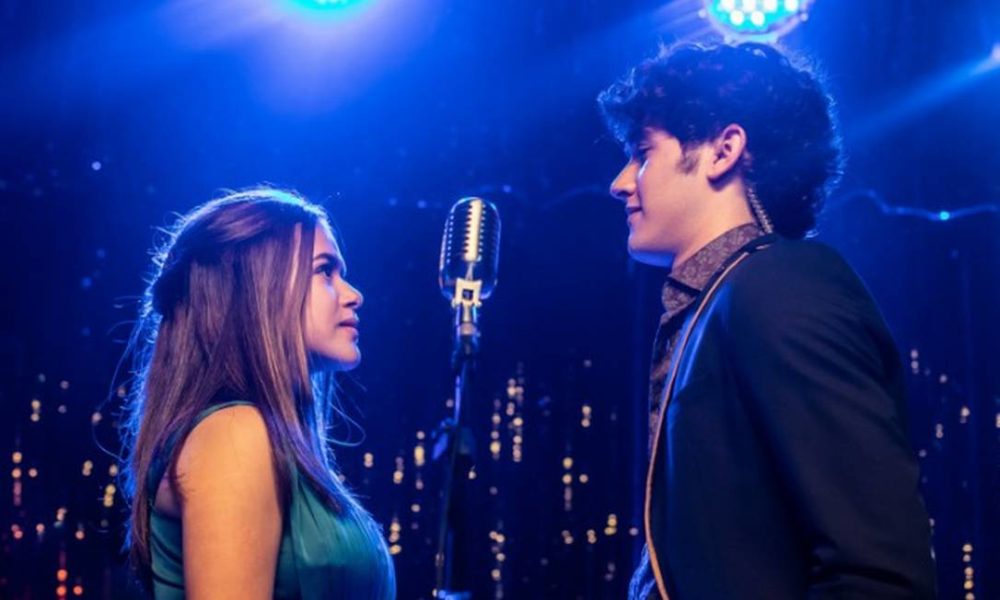 Cena romântica com um casal cantando juntos em um palco com iluminação azul, refletindo uma conexão especial entre eles.