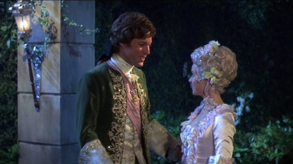 Cena de um filme clássico com um casal vestido com trajes elegantes no jardim à noite, destacando o charme e elegância da época.