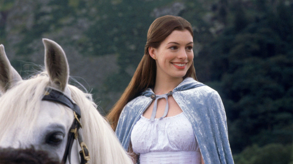 Mulher sorridente com capa azul e vestido branco ao lado de um cavalo branco em meio à natureza, representando fantasia e elegância.