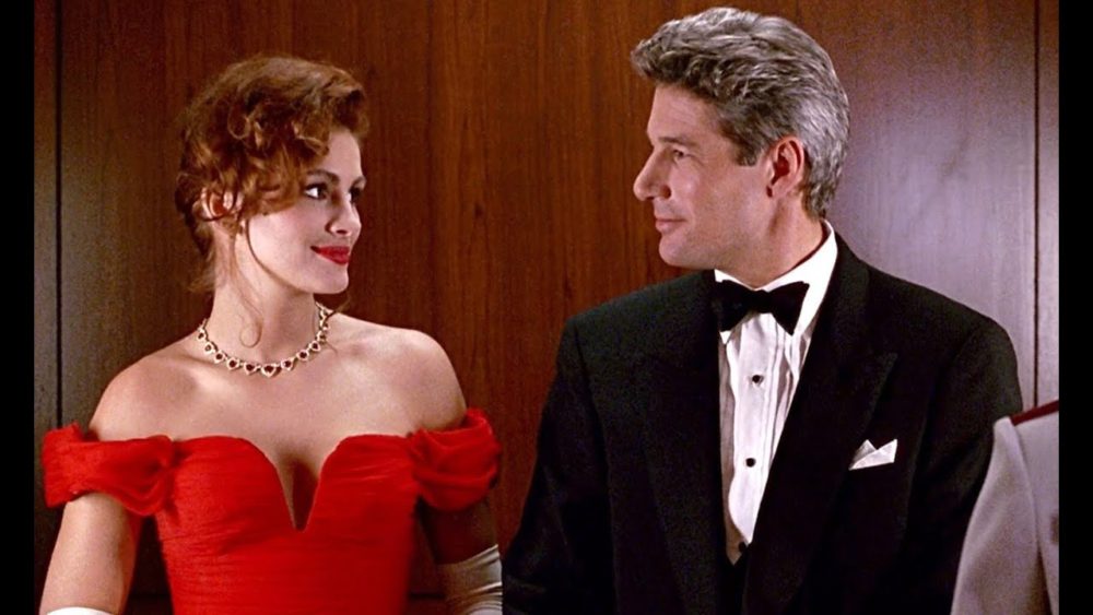 Cena de um filme clássico com uma mulher com cabelo ruivo e vestido vermelho elegante e um homem com bochechas grisalhas e terno preto em conversa em um ambiente sofisticado.