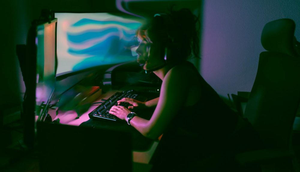 Pessoa trabalhando em computador com teclado iluminado em ambiente de estúdio, com luzes coloridas e monitor ao fundo, aparência de trabalho criativo à noite
